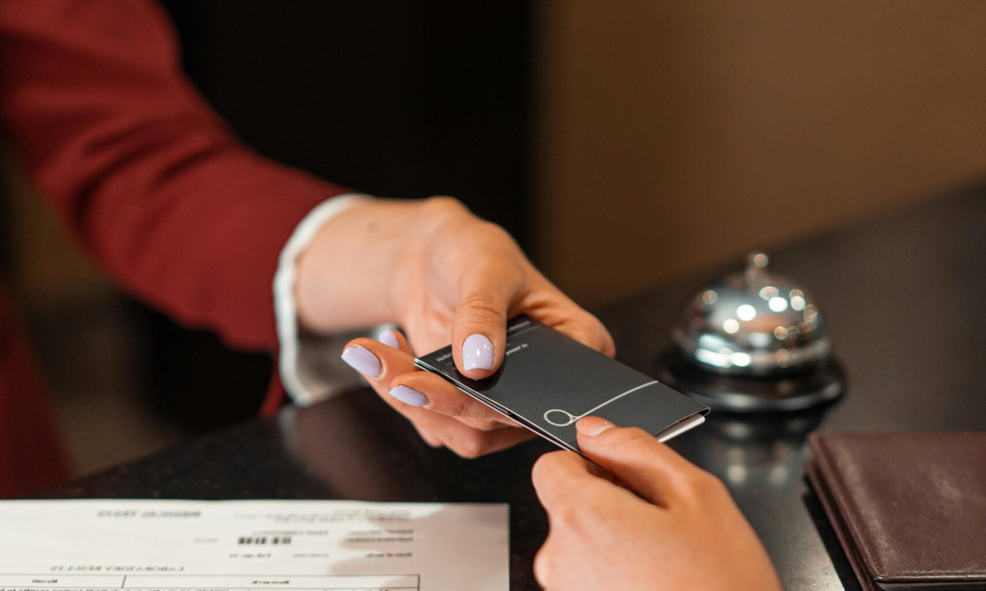 A hotel receptionist hands a key card to a guest over the counter, with documents visible.